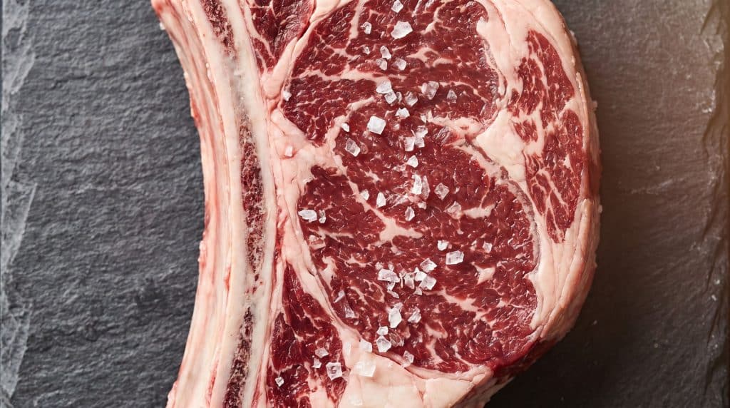 Cattleman's Cutlet: The Mini Roast Beef Meal for One 1 Close-up of a raw cattleman's cutlet showing the rib bone and rich intramuscular marbling through the longissimus dorsi muscle on a slate board.