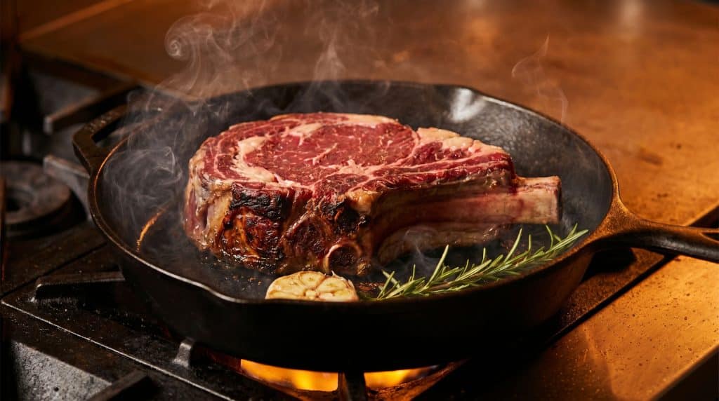 Cattleman's Cutlet: The Mini Roast Beef Meal for One 7 A thick cattleman's cutlet searing in a cast iron pan over high heat, with a deep brown Maillard crust forming and wisps of smoke rising from the hot surface.